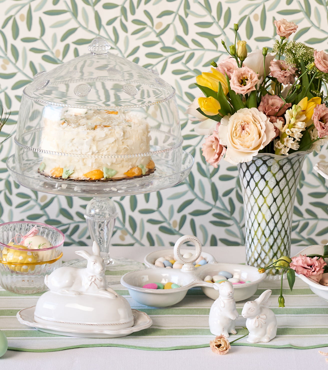 Easter-themed table setting with cake, flowers, and decorative items against a leafy green wallpaper.