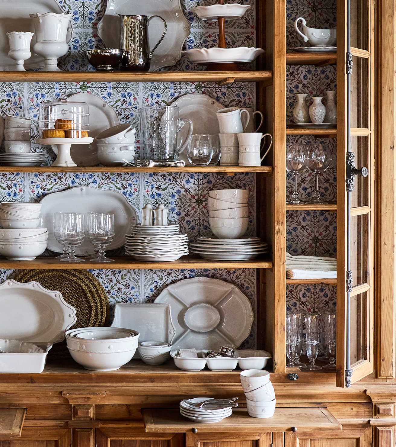 Wooden cabinet with shelves displaying various ceramic and glass dishes against a patterned wall.