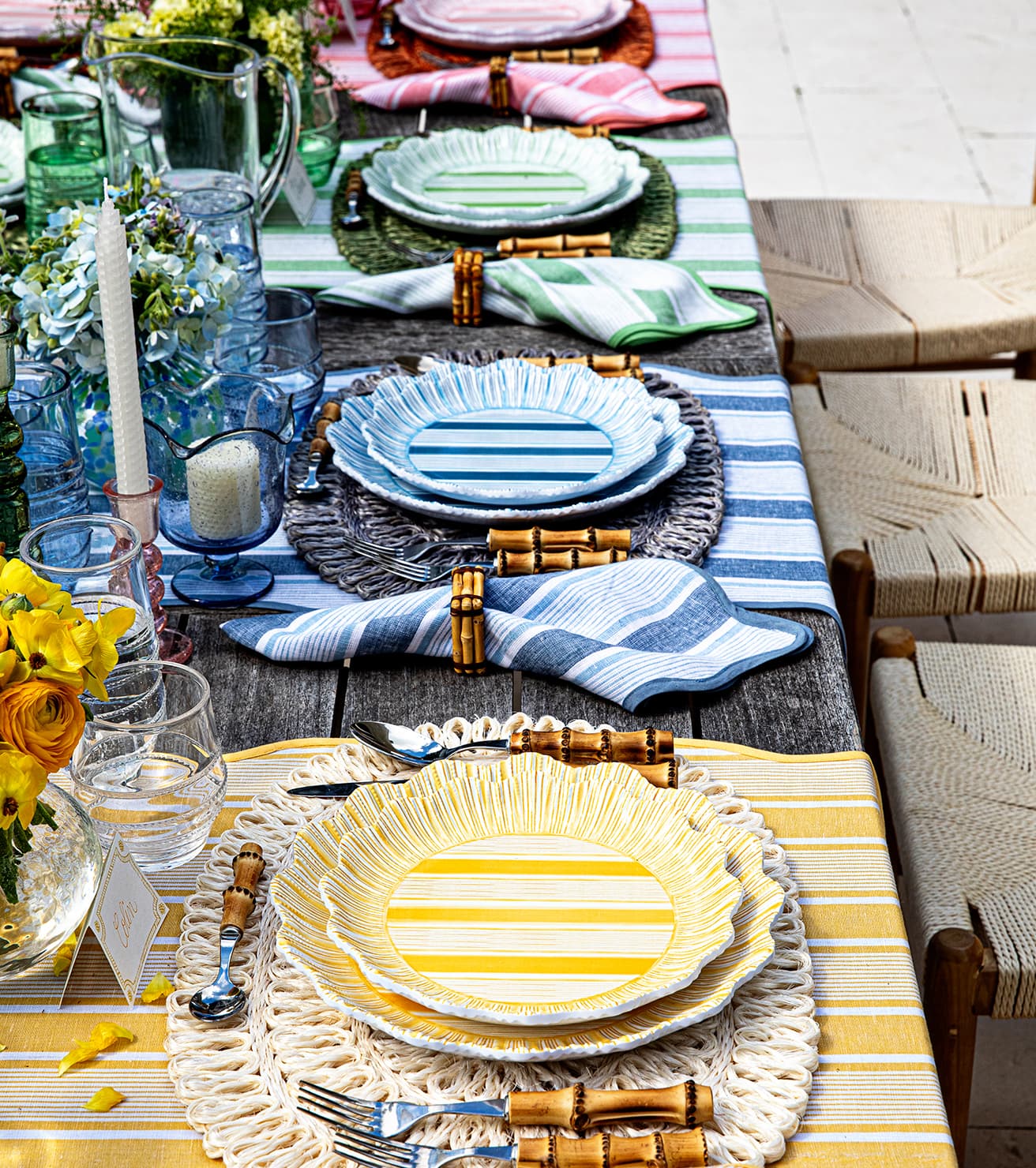 A long outdoor dining table is set with colorful striped plates, bamboo-handled cutlery, and striped napkins.