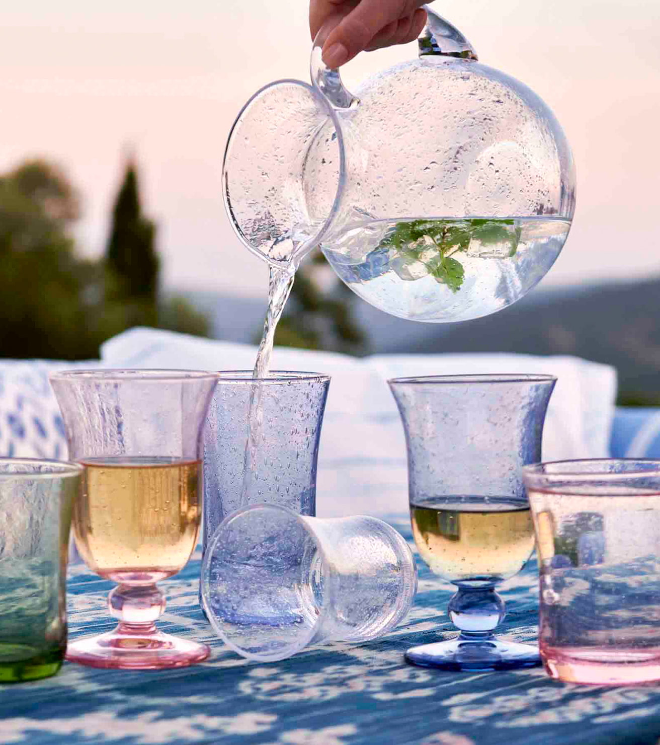 Clear glass pitcher being filled with water, surrounded by various glasses on a table outdoors.