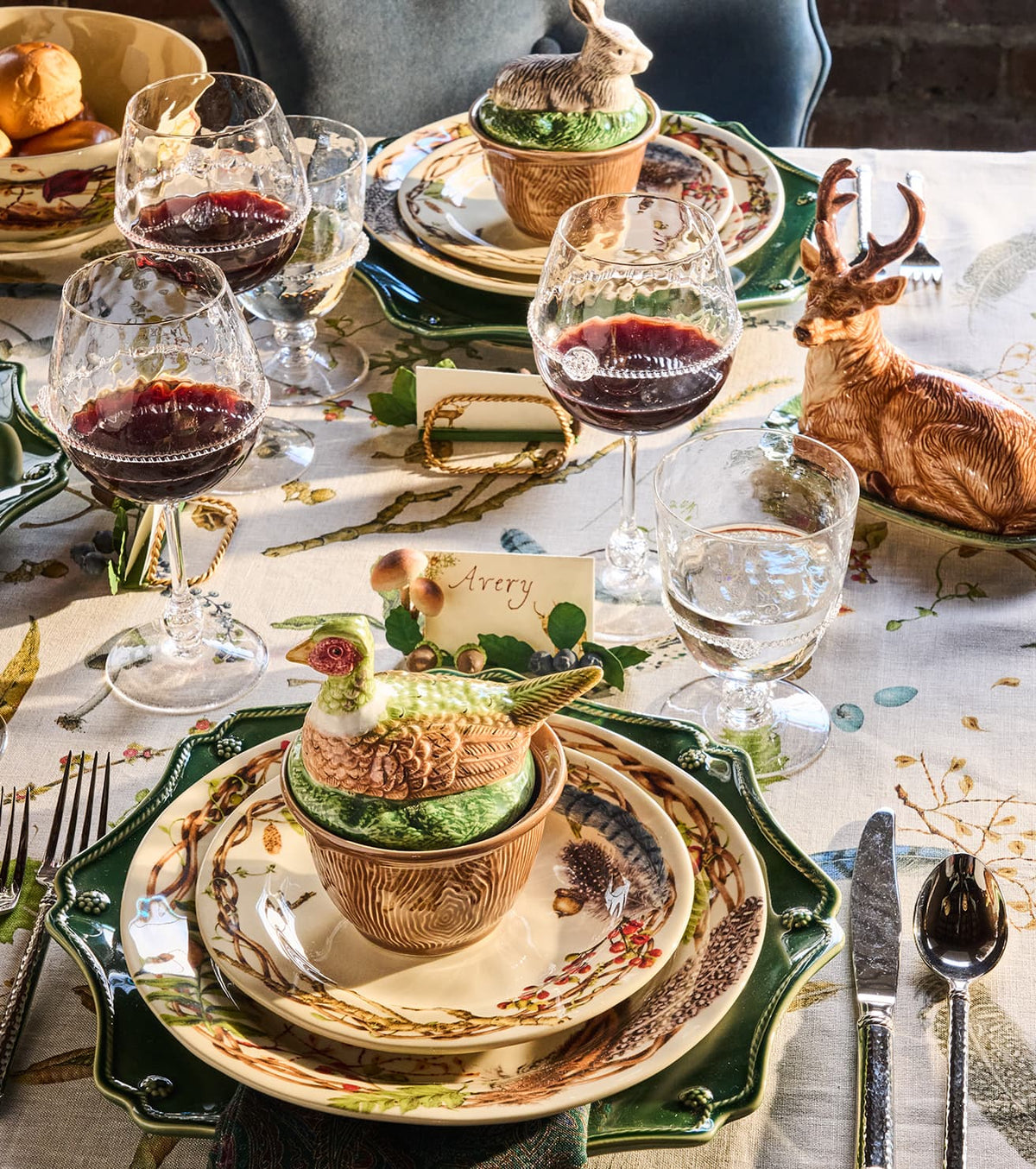 A styled dining table setting with layered ceramic plates featuring a woodland deer and leaf pattern, wine glasses, a green duck-shaped centerpiece in a ceramic cup, and a deer figurine on a tablecloth with botanical print.