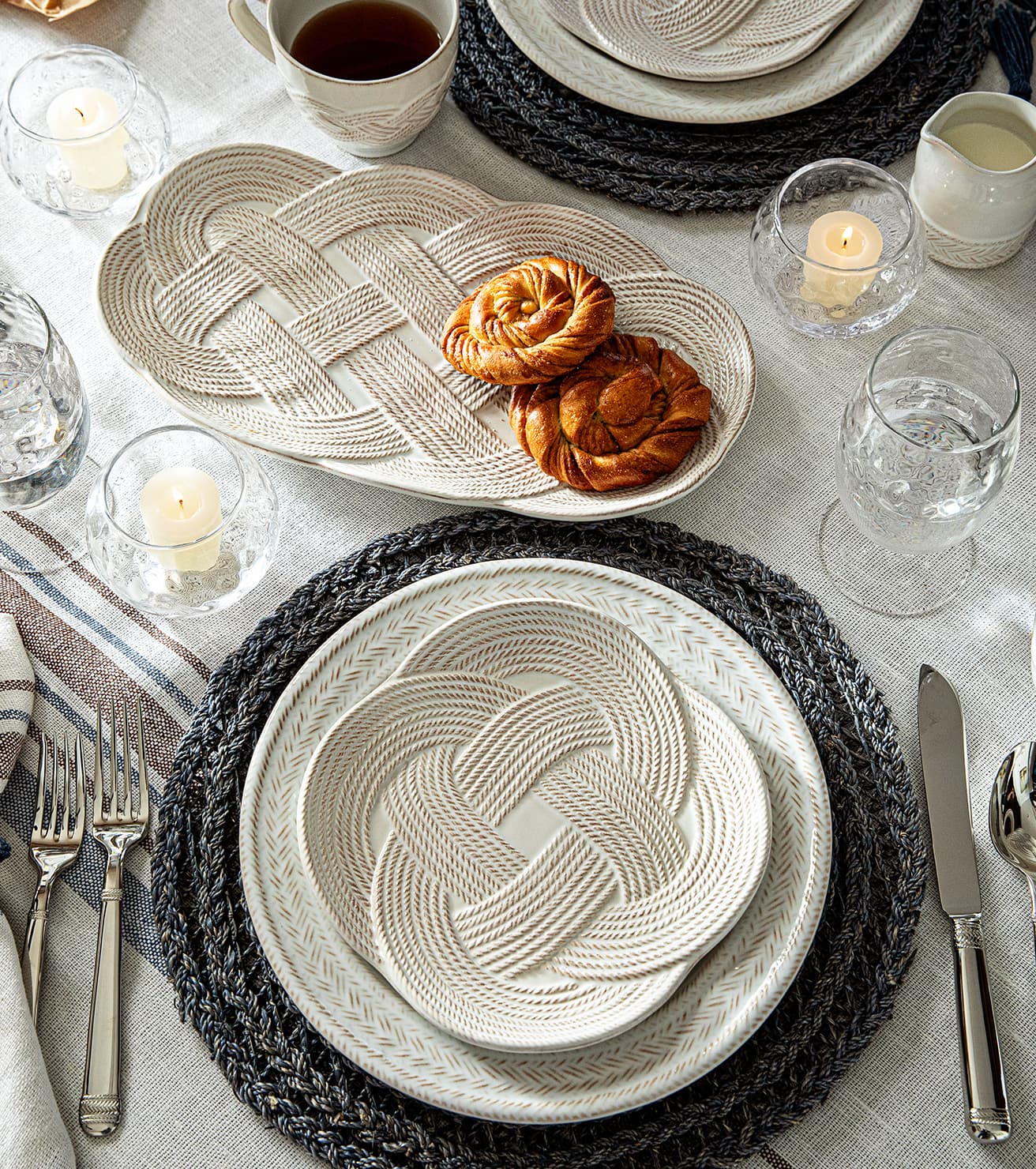 Elegant table setting with woven plates, candles, and cutlery on a light surface.