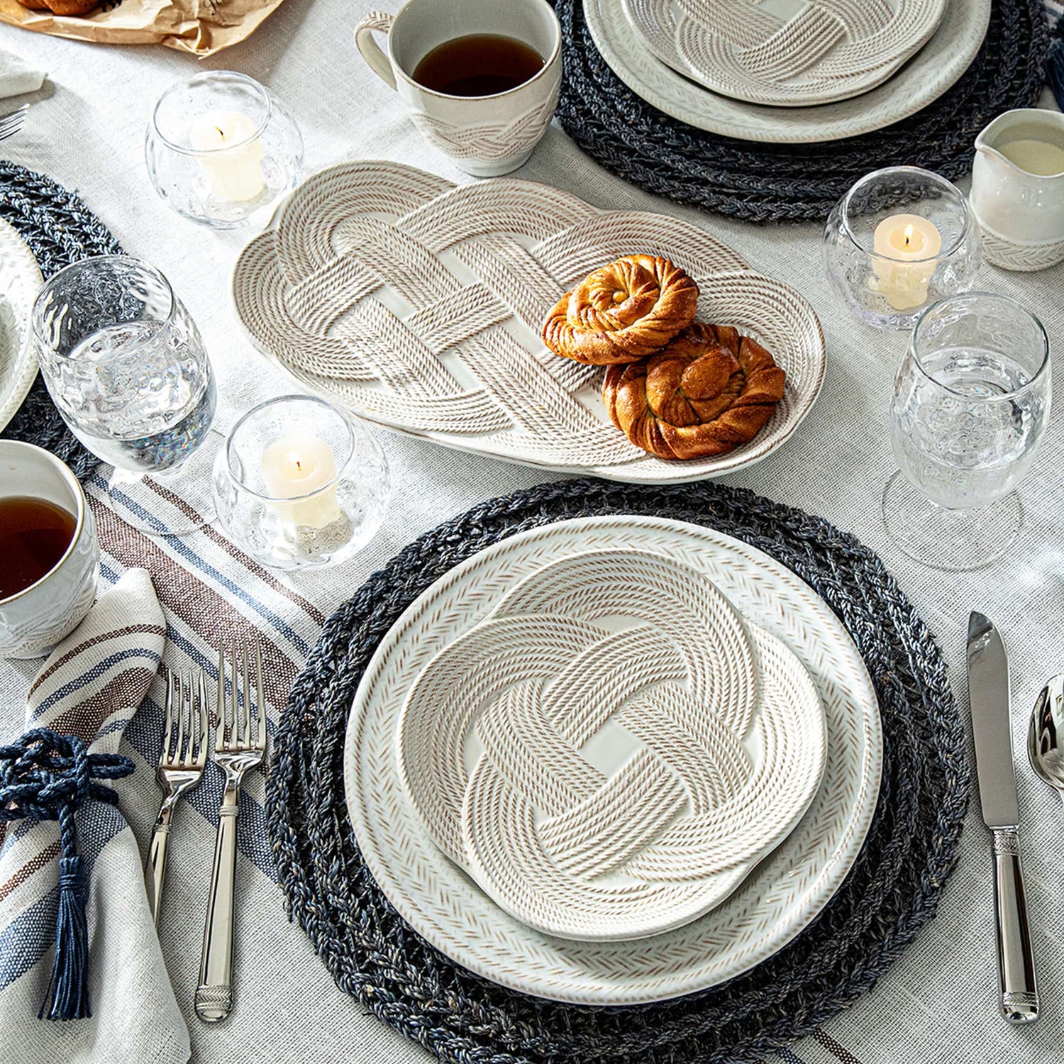 A styled table setting featuring a coordinated set of textured ceramic plates with a braided knot pattern, dark gray woven placemats, silver cutlery, and clear glass candleholders on a light linen tablecloth.
