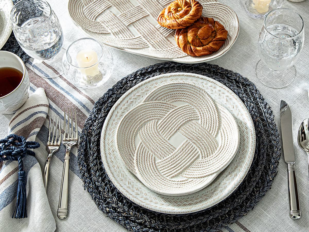 Table setting with woven plates, cutlery, and glasses on a textured tablecloth.