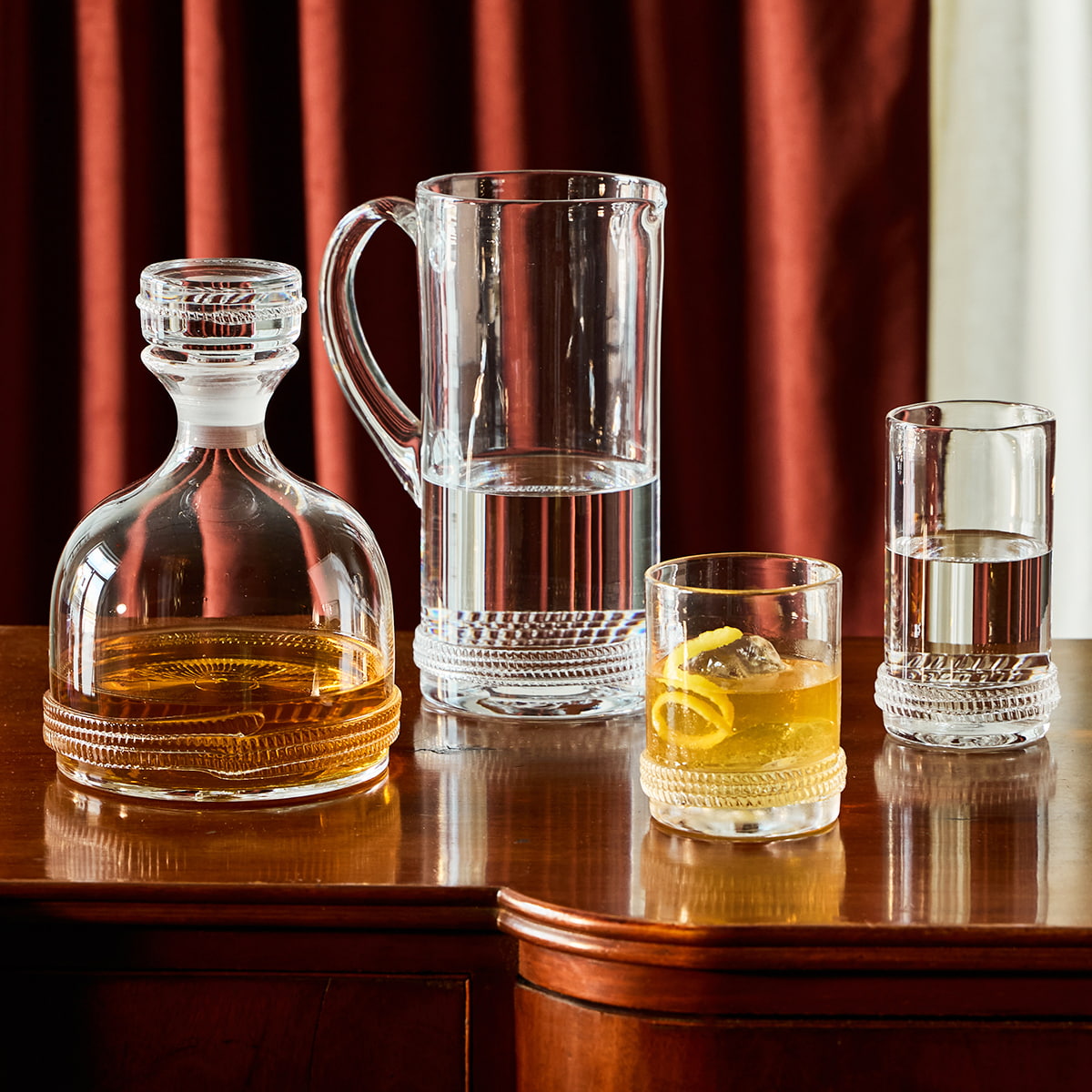Set of glass decanter, pitcher, and glasses from the dean collection on a wooden surface with a red curtain background.