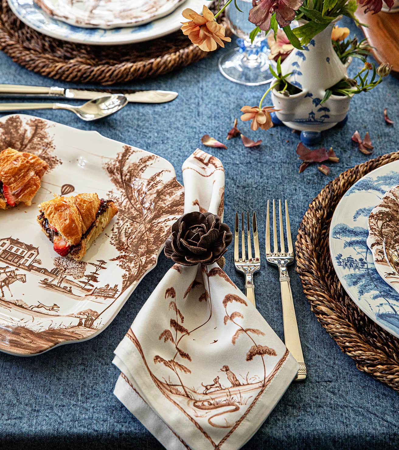 A styled table setting featuring a cream napkin with a brown toile-style landscape print and a dark fabric rose napkin ring, paired with silver forks on a blue tablecloth. Matching patterned plates and woven placemats surround the setting.