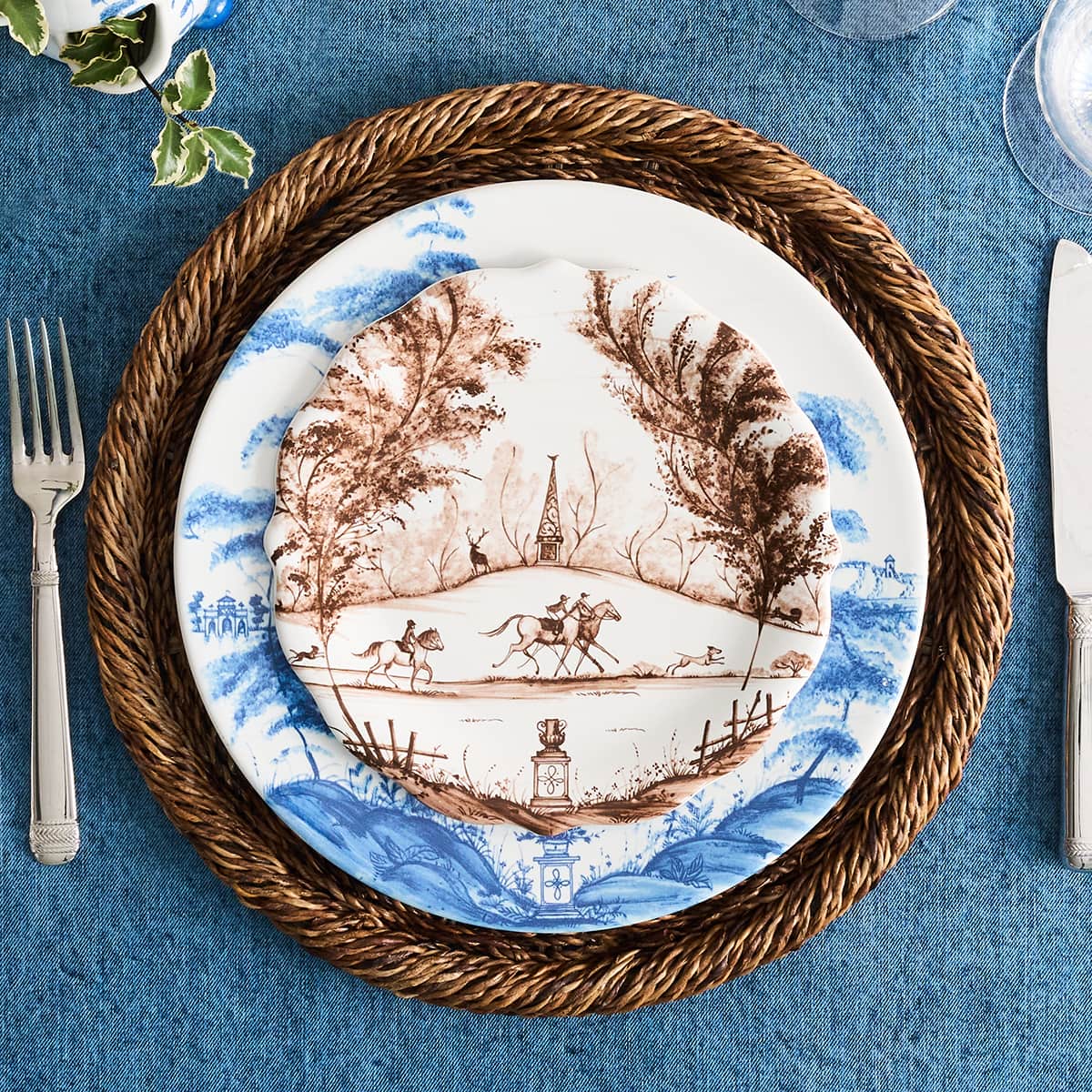 A place setting featuring two stacked ceramic plates with a classic blue and brown toile-style landscape print, layered on a round braided brown placemat over a textured blue tablecloth, with silver cutlery visible on the sides.