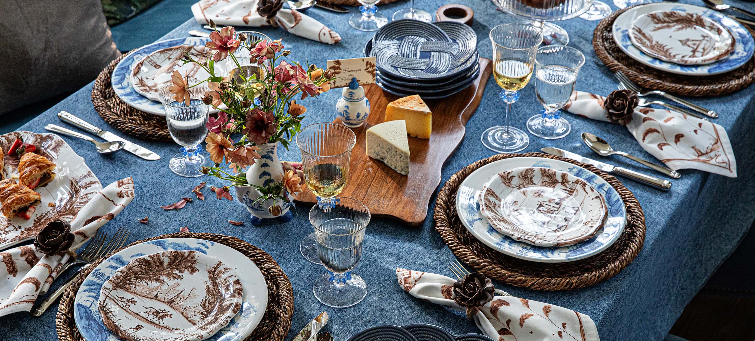 An elegantly set dining table featuring a blue tablecloth, blue and white patterned plates, woven placemats, clear glass goblets, and a central floral arrangement with cheese on a wooden board.