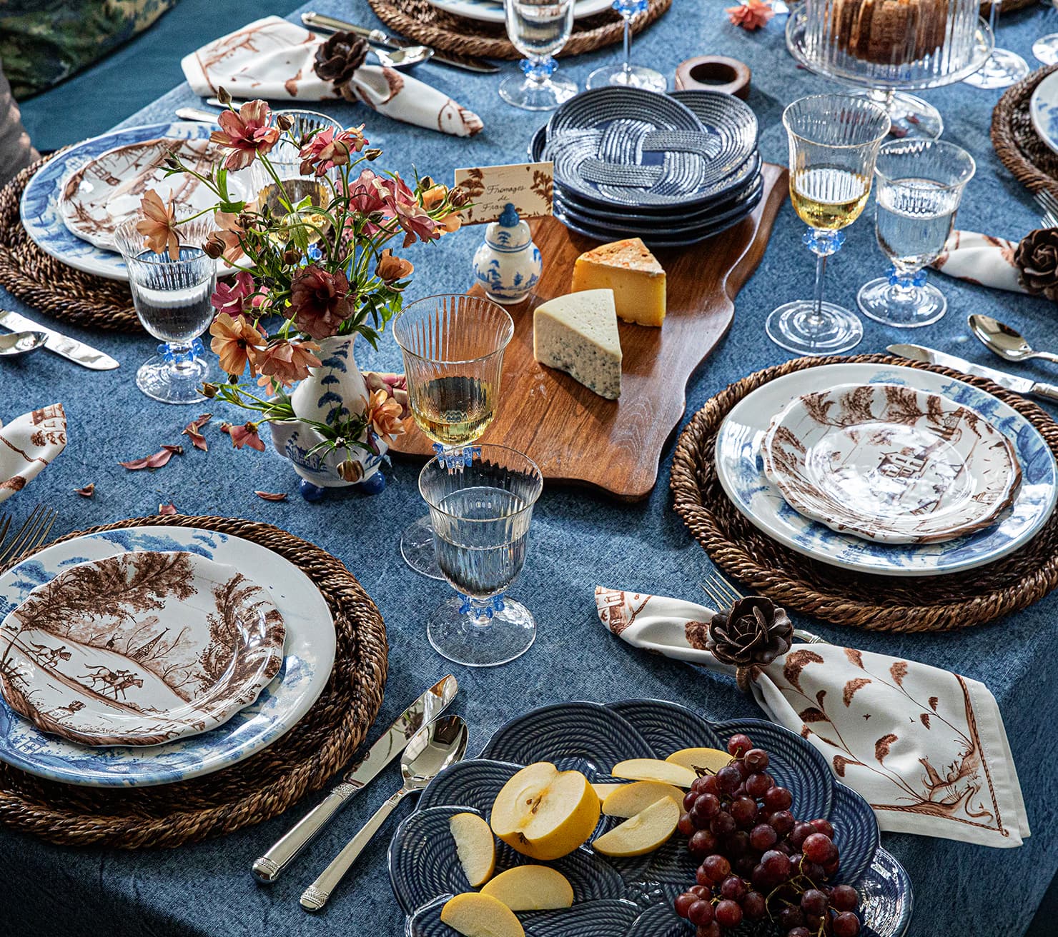 An elegantly set dining table featuring blue and white patterned plates, woven placemats, clear glass goblets, a floral centerpiece, cheese, grapes, and apple slices on a blue tablecloth.