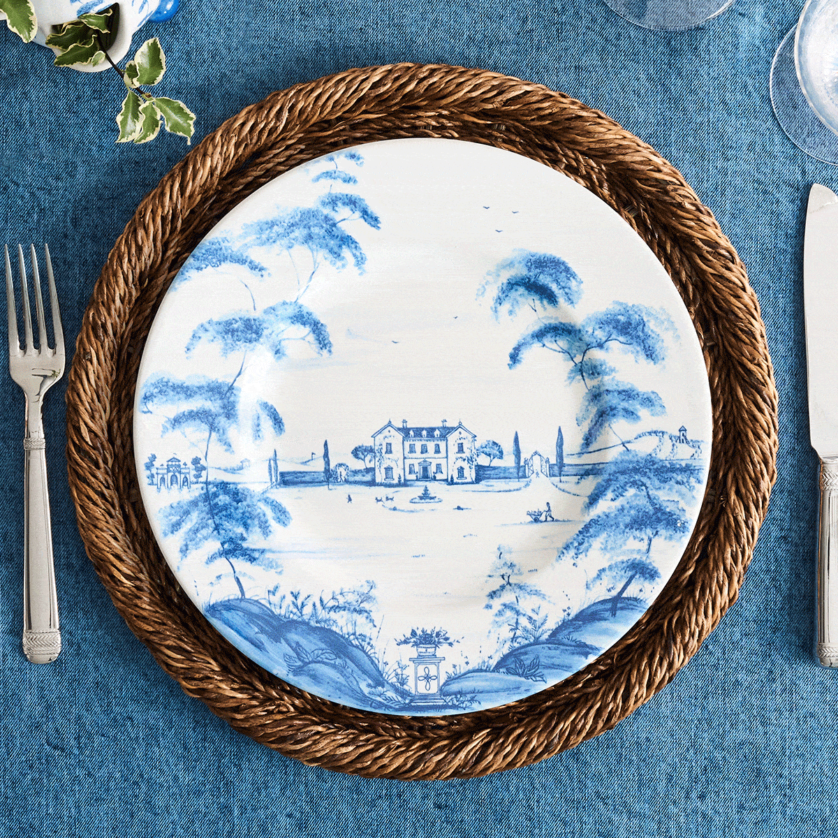A moveable gif place setting with a blue and white landscape plate, woven charger, silver cutlery, a napkin with a brown napkin ring, and a blue tablecloth.
