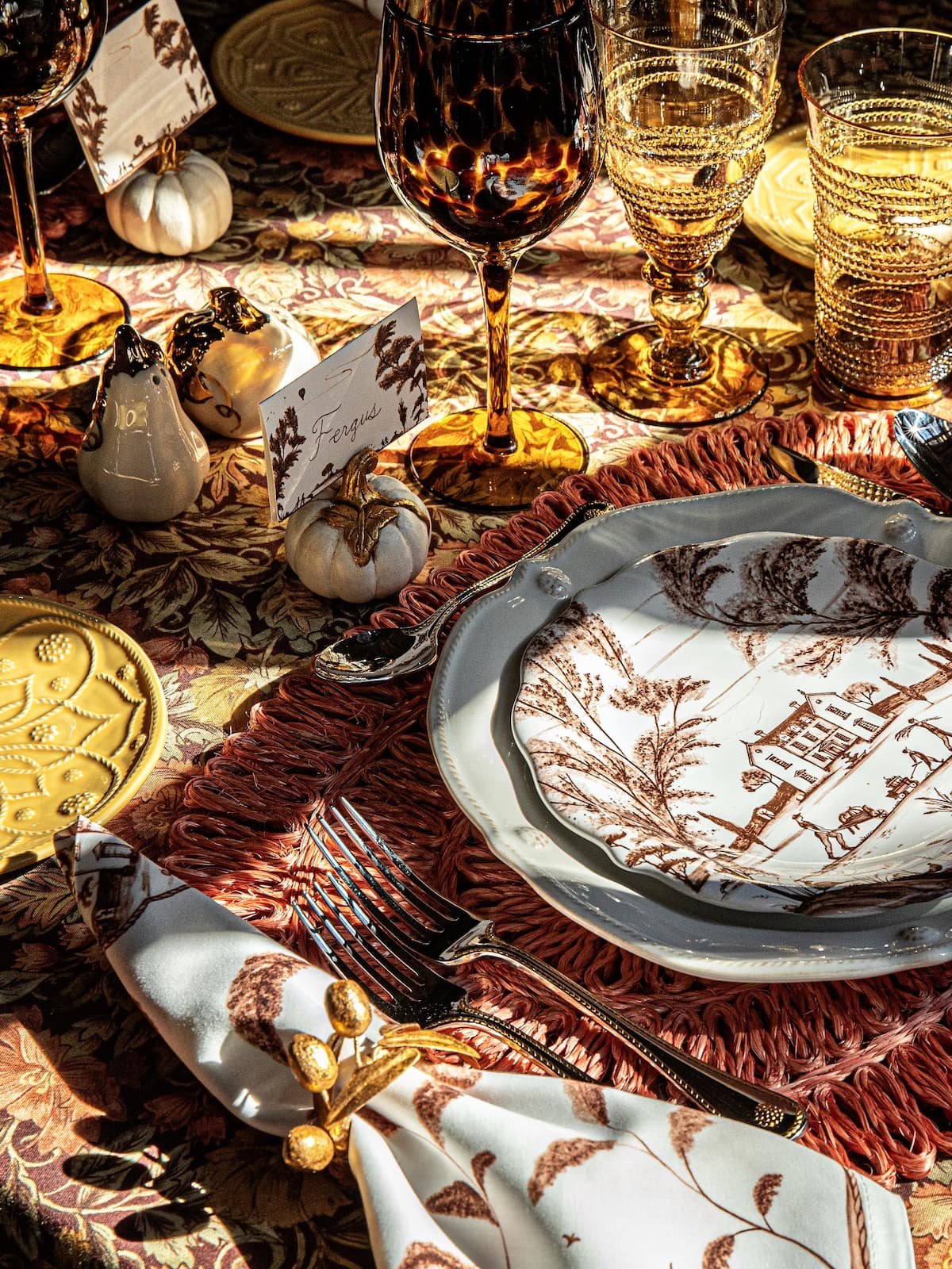 Decorative table setting with wine glasses, plates, and pumpkins on a patterned tablecloth.