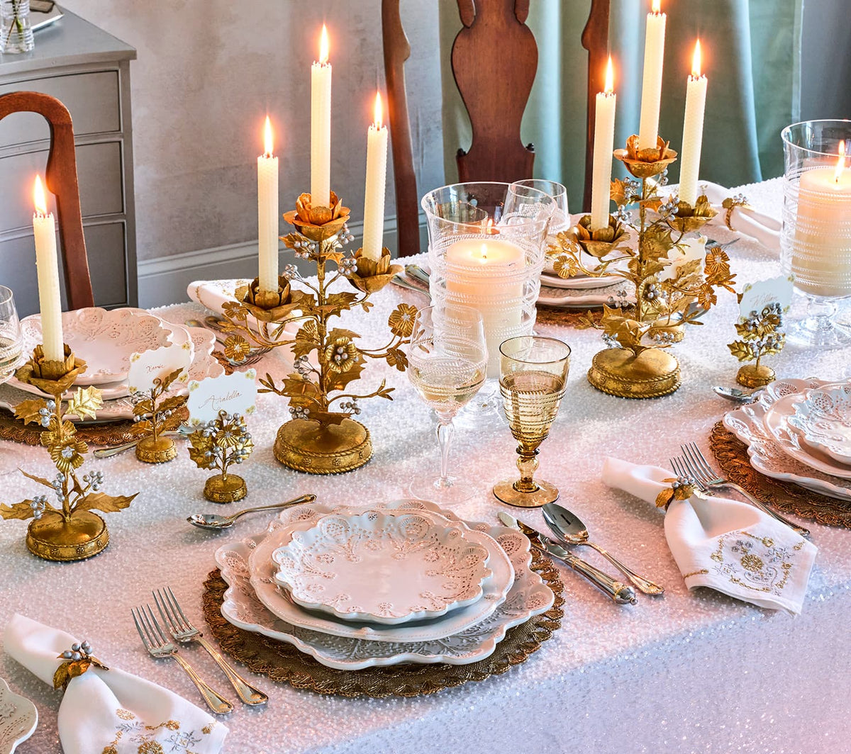 An ornate gold and white table setting with multiple candlesticks, plates, napkins, and goblets arranged on a table covered with a white tablecloth. The scene is styled for a formal dinner table with lit taper candles and reflective metallic accents.