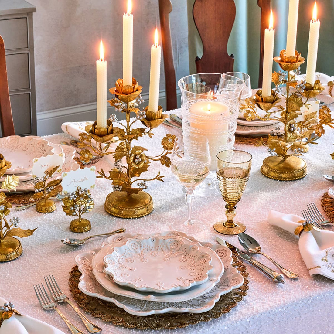 An ornate gold and white table setting with multiple lit taper candles, clear and gold goblets, white plates with scalloped edges, and decorative gold floral candle holders on a white tablecloth.