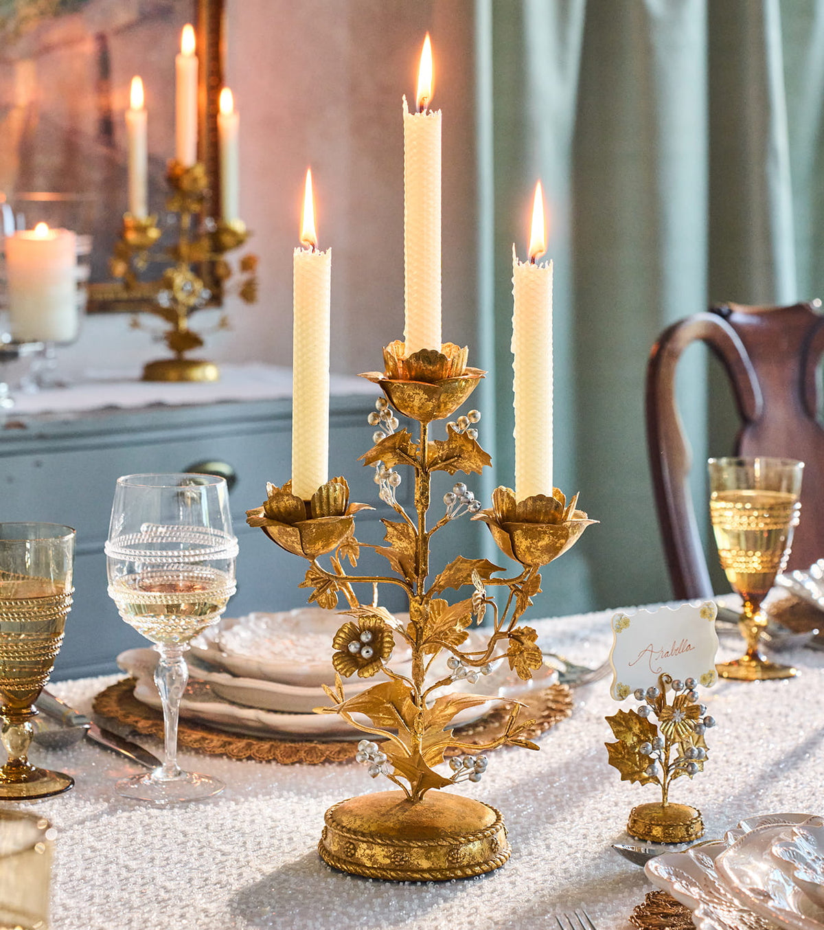 An ornate gold floral candle holder centerpiece with three lit taper candles on a decorated dining table, surrounded by glassware, folded white napkins, and a place card.