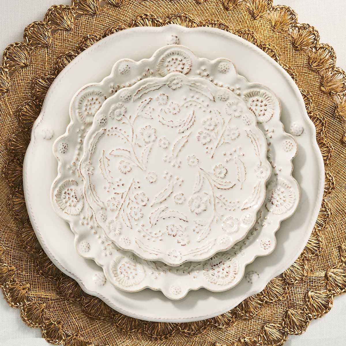 Three stacked ceramic plates with scalloped edges and raised floral relief patterns, shown on a textured gold placemat.