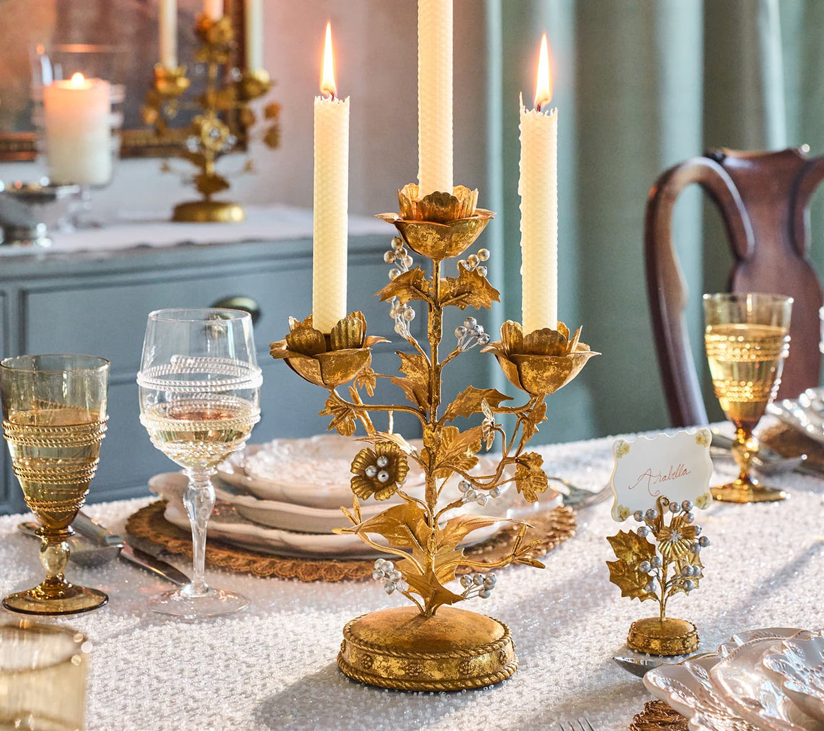 An ornate gold metal candlestick centerpiece with floral details and lit taper candles, styled on a decorated dining table with glassware, plates, and a small matching place card holder.