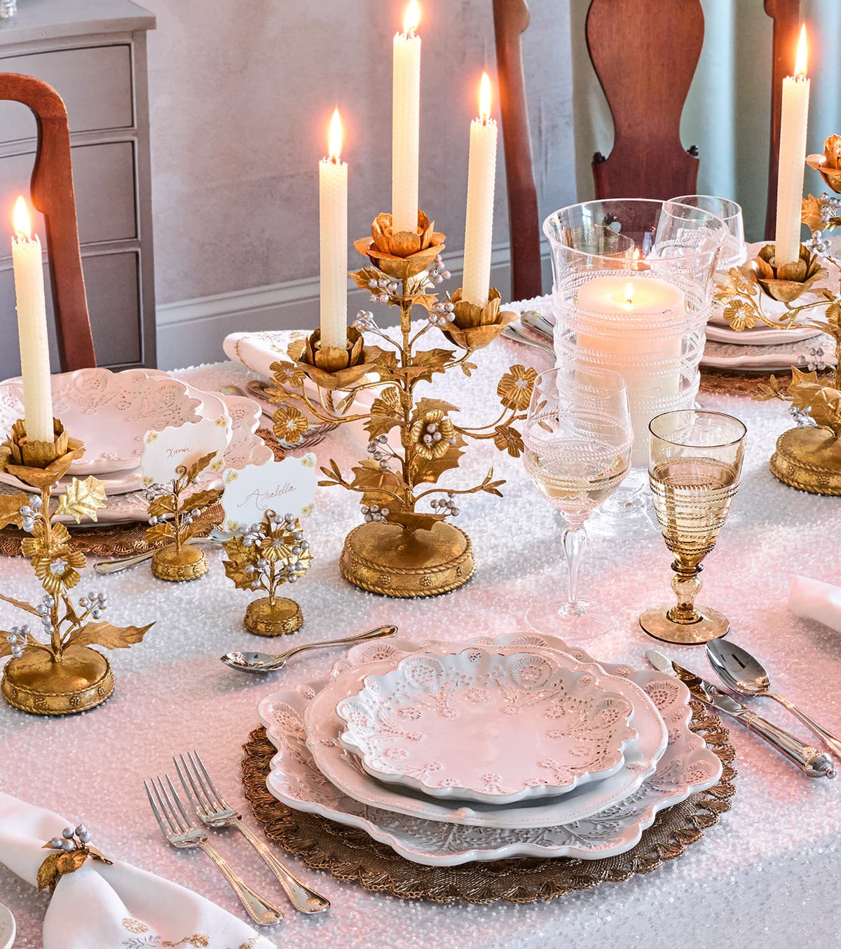 An elegant table setting featuring ornate gold floral candlesticks with lit taper candles, matching gold and white plates, clear glass goblets, and white napkins on a textured white tablecloth.