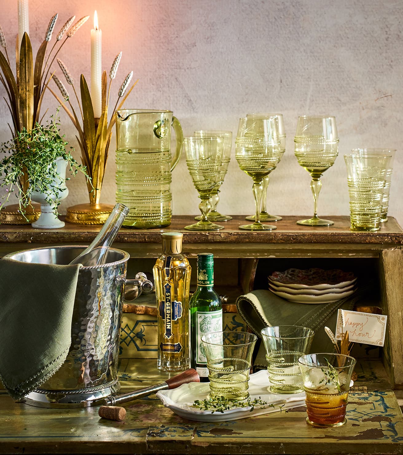 A styled bar and dining scene with multiple ribbed glassware pieces, including a pale green pitcher, four wine glasses, and two matching tumblers, arranged on rustic wooden shelves alongside bottles, an ice bucket, candles, and decorative wheat stems.