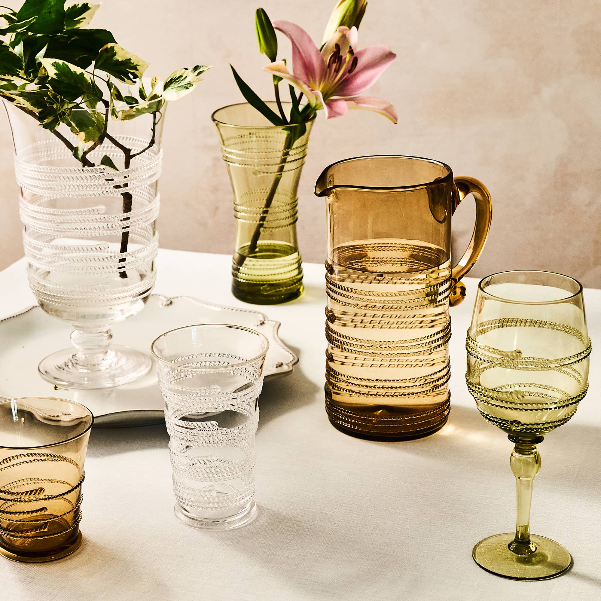 A set of ribbed glass tableware in clear and amber tones, arranged on a light tabletop with decorative vases, flowers, and a plant. The set includes a large amber pitcher, a wine glass, a tumbler, a stemware glass, and a pedestal plate.