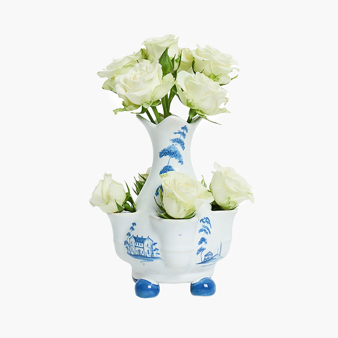A white ceramic three-bowl flower vase with blue hand-painted landscape motifs and blue feet, filled with white roses, on a light gray background.
