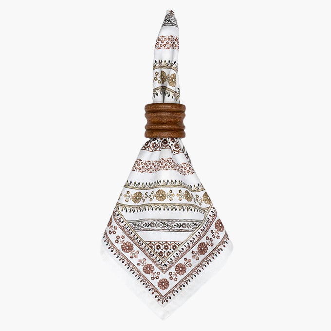 A white napkin with intricate brown, beige, and black geometric and floral patterns, tied around a brown wooden napkin ring on a light gray background.