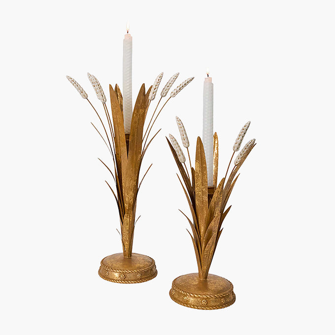 A pair of gold-toned metal candlestick holders designed to look like wheat stalks, each holding a white taper candle, set against a plain white background.