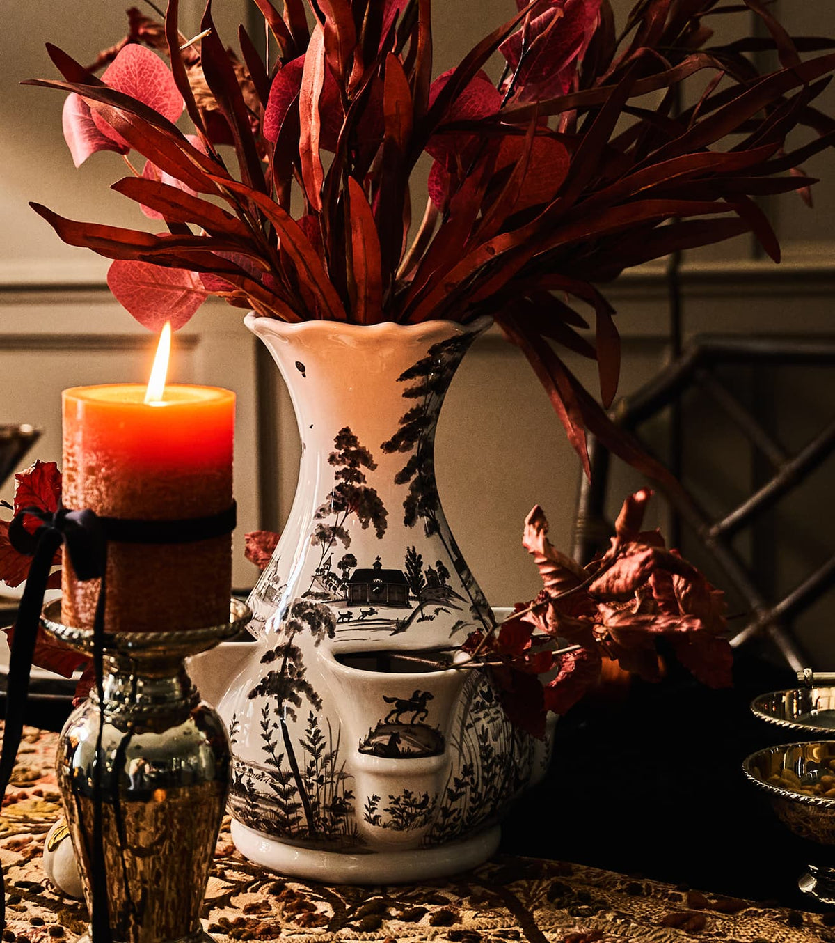 A white ceramic oil burner with black landscape illustrations, styled with a lit orange candle, dried red foliage, and a rustic table setting.