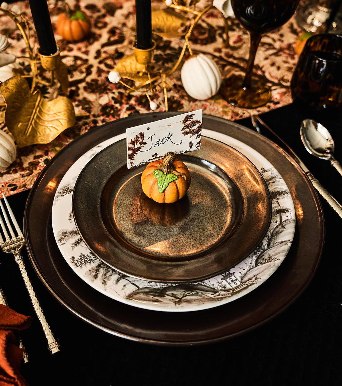 A styled autumn table setting featuring a place setting with a dark brown charger plate, a white plate with gray leaf patterns, a dark bronze plate, and a small orange pumpkin decoration with a place card on top.