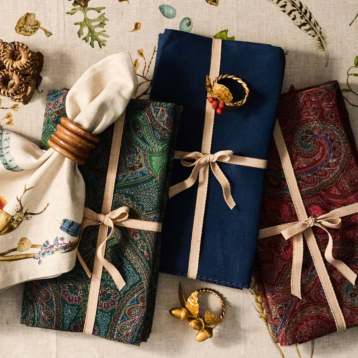 Three folded patterned textiles tied with beige ribbon are arranged on a light fabric surface with decorative botanical and wood accents. The textiles feature ornate paisley and floral prints in navy, teal, and burgundy tones, with one cream textile showing deer and floral motifs.