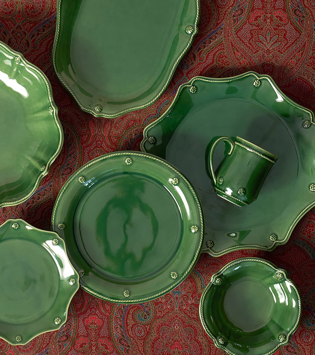 A coordinated green ceramic tableware set arranged on a red patterned fabric background, including plates, a bowl, a mug, and serving platters with decorative raised edges and small metallic accents.