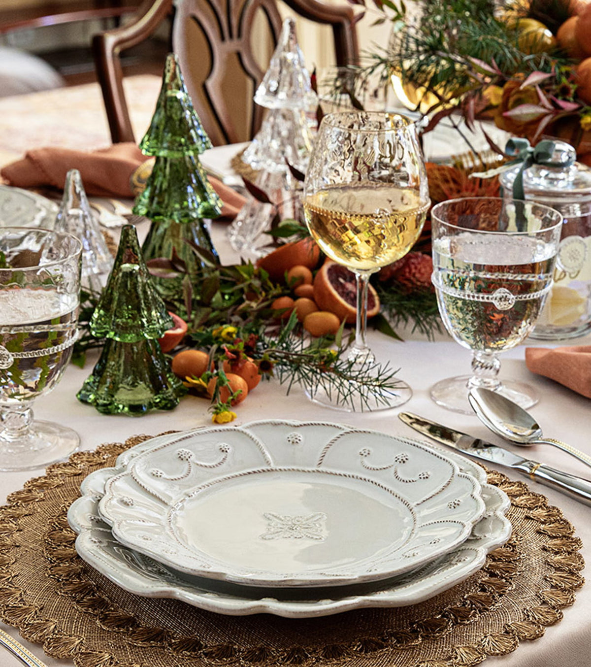A styled holiday table setting featuring a stack of white ceramic plates with scalloped edges and a subtle star motif in the center, set on a woven placemat with wine glasses, green glass trees, and autumn decorations.