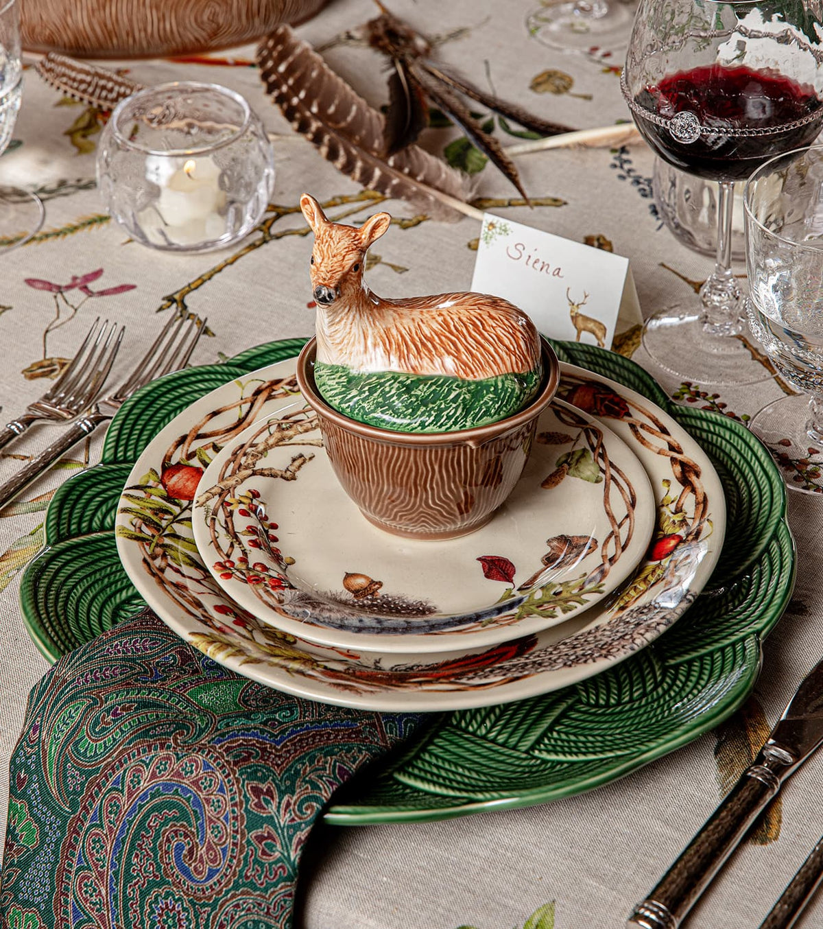 Table setting with layered plates, a green napkin, animal-themed bowl with a deer lid, wine glasses, silverware, and a place card reading “Siena” on a nature-print tablecloth.