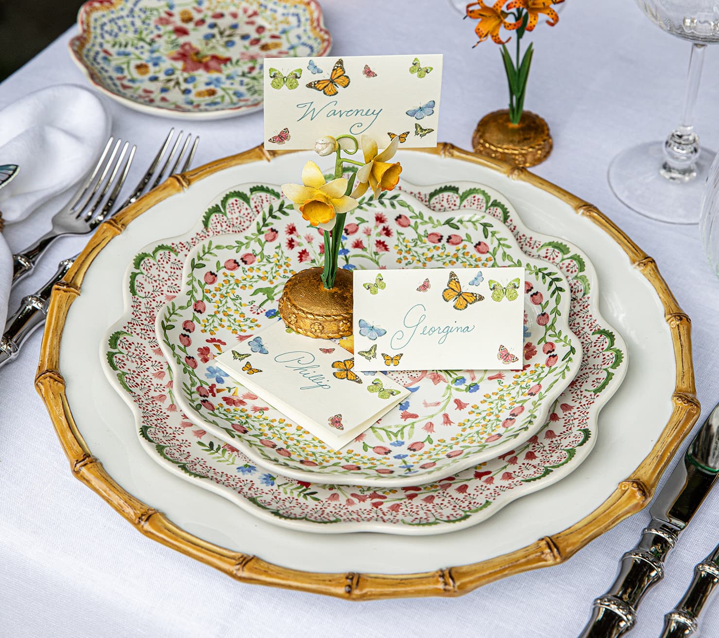 Decorative table setting with floral plates, cutlery, and name cards on a white tablecloth.