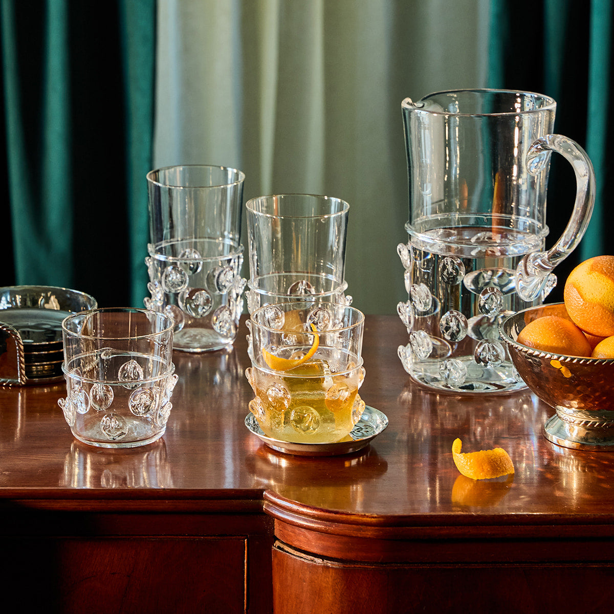 Set of glass tumblers and a pitcher on a wooden table with oranges.