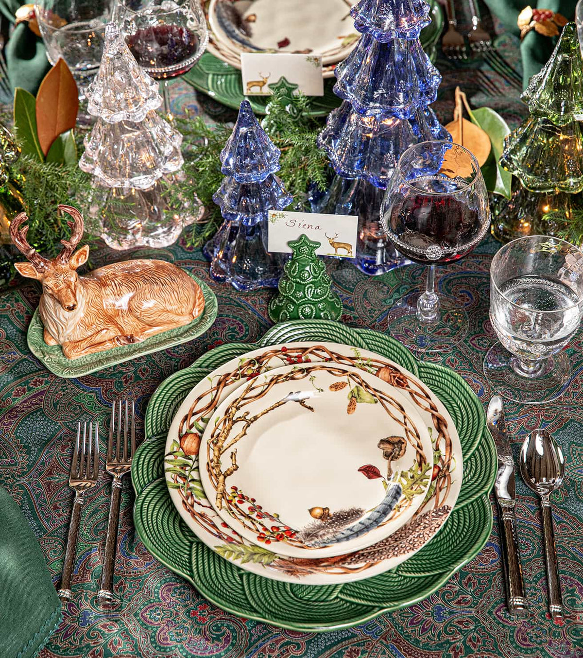 A festive holiday table setting featuring green ceramic plates with a tree branch and bird pattern, silver cutlery, wine glasses, and decorative glass Christmas trees on a patterned tablecloth.