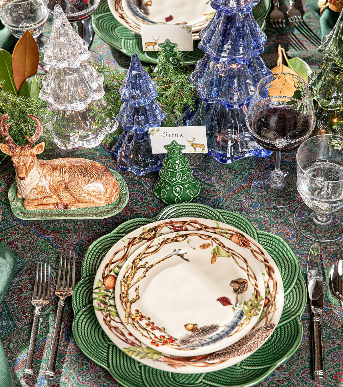 A styled holiday table setting featuring a green scalloped charger plate under stacked white plates with a woodland wreath border, alongside green and blue glass tree ornaments, a ceramic deer centerpiece, and patterned table linens.