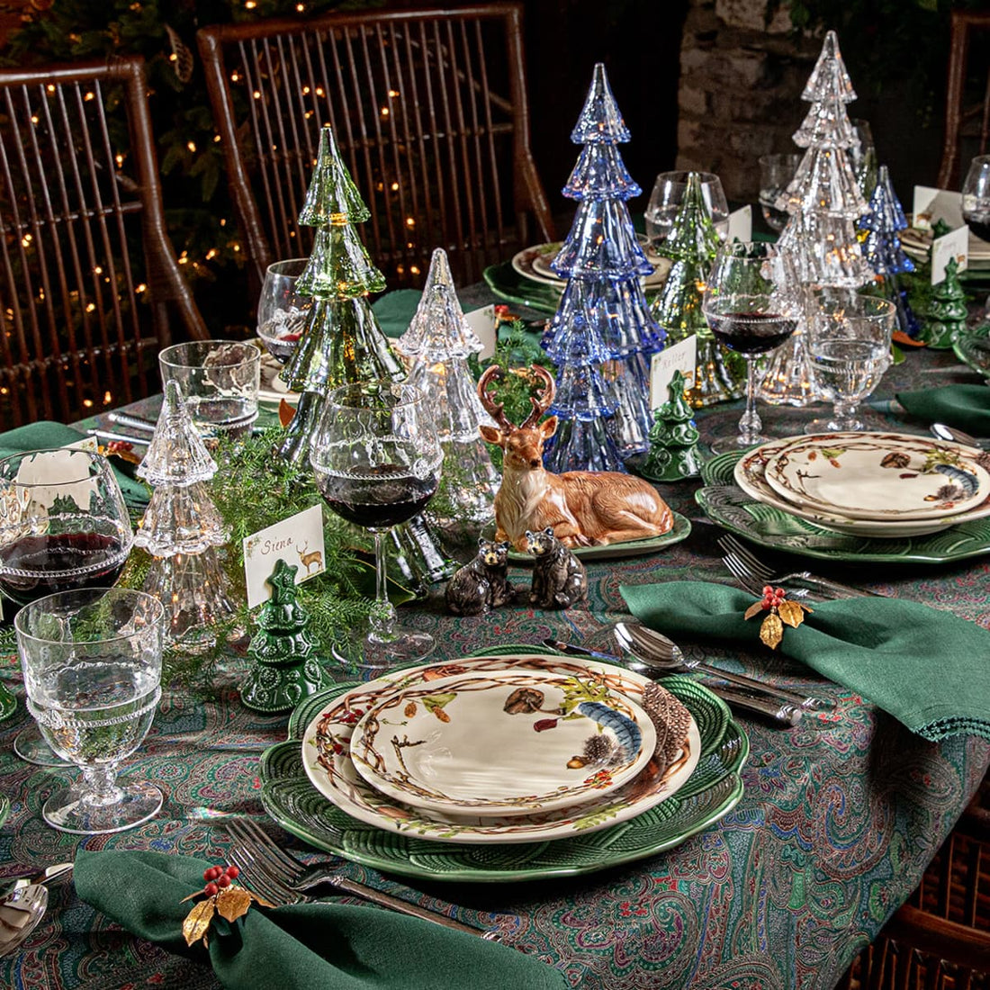A festive holiday table setting with green and clear glass Christmas tree-shaped drinking glasses, layered plates, green napkins, and small decorative figurines, including a deer and miniature trees, arranged on a patterned tablecloth.