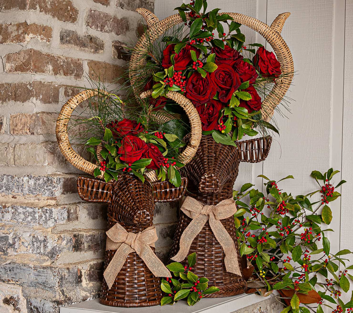 Two wicker reindeer-shaped decorative planters with large curved horns, tied with beige burlap bows, filled with red roses, green foliage, and red berries, displayed against a brick and white door background.