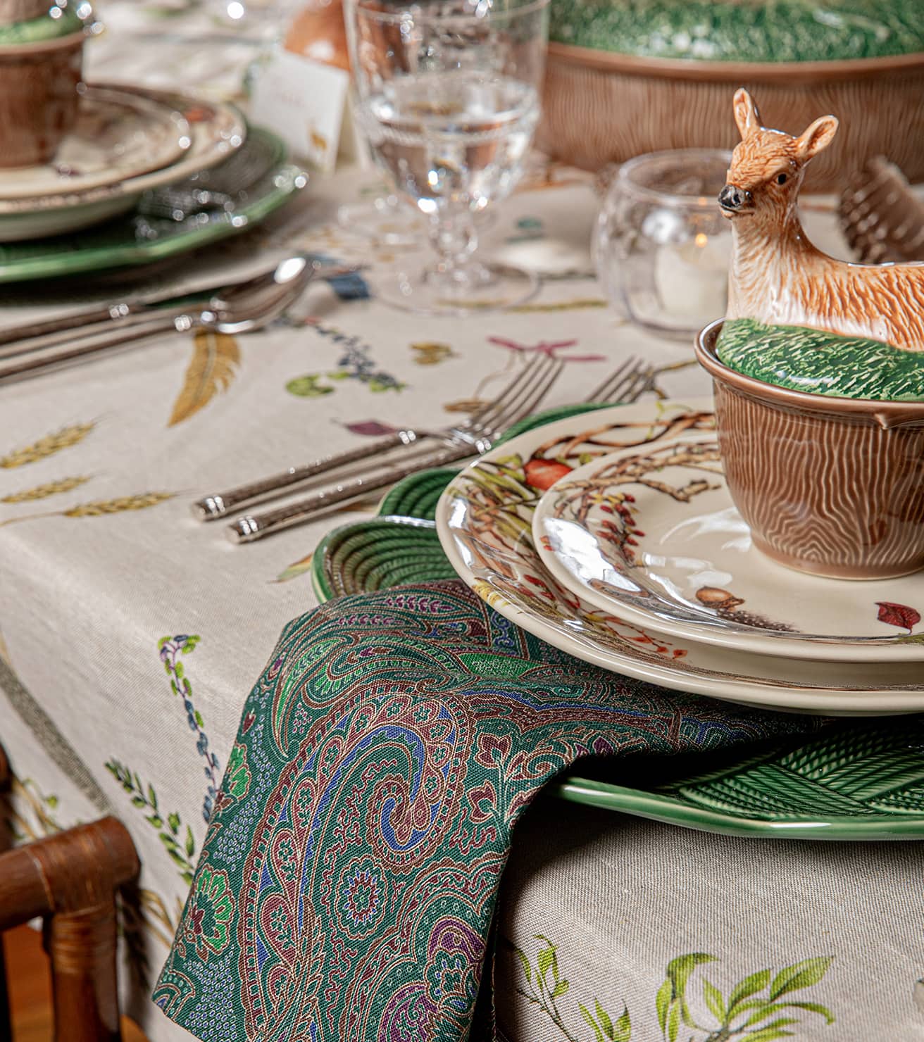 A dining table set with patterned plates, green napkins, glassware, and a bowl with a deer figurine lid on a nature-themed tablecloth.