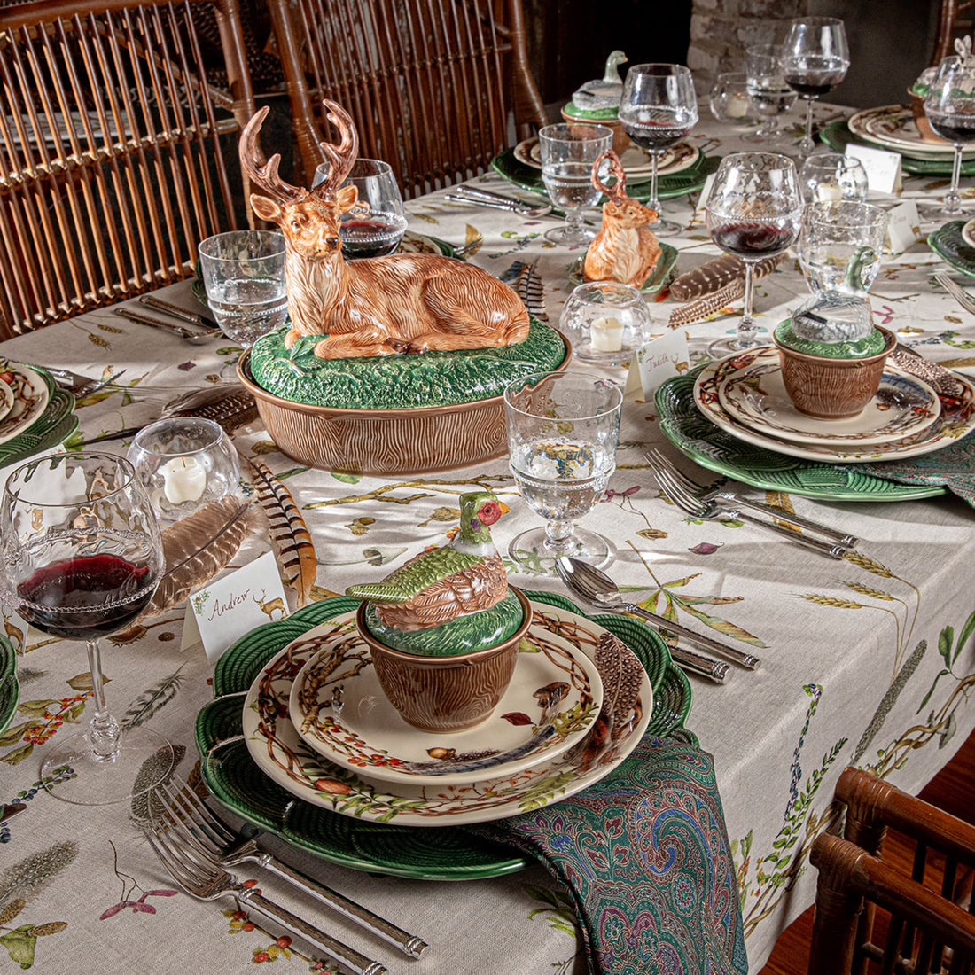 A rustic, cabin-style dinner table fully set with a beige tablecloth featuring green and brown woodland patterns, matching ceramic plates, wine glasses, and a central decorative ceramic deer casserole dish as centerpiece.