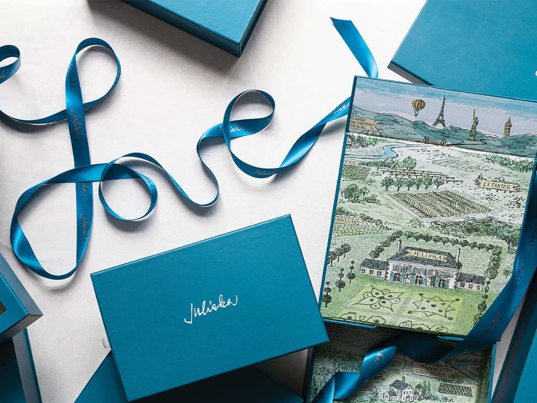 A luxury gift set featuring a teal blue box with the script brand name 'Juliska' on the lid, another box showing a detailed landscape illustration of a French chateau, and matching teal ribbons, all arranged on a white background.