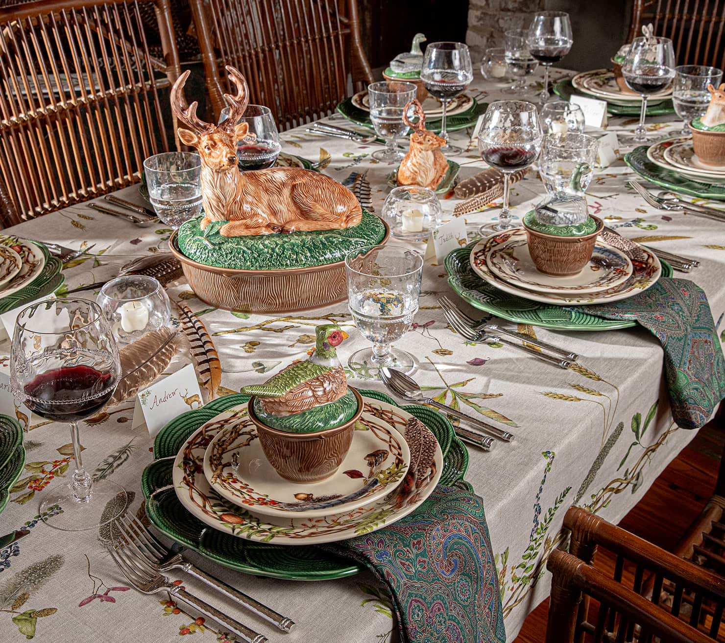 A styled dining table set with a beige floral tablecloth, green ceramic plates, matching napkins, crystal glasses, and decorative ceramic deer figurines on top.