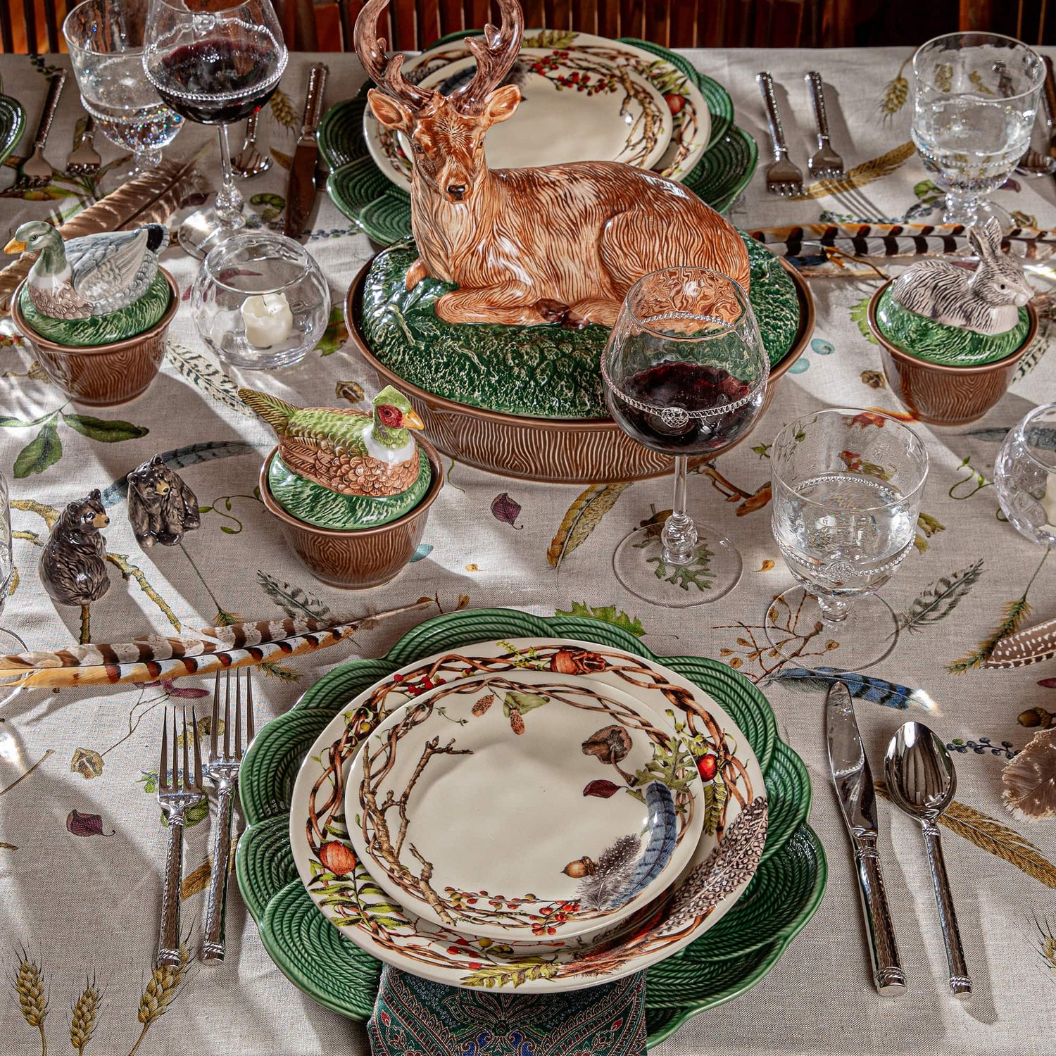 A styled dining table setting featuring a rustic woodland theme with a ceramic deer centerpiece, matching green and cream patterned plates, silver flatware, wine glasses, and decorative bird and feather accents on a patterned tablecloth.