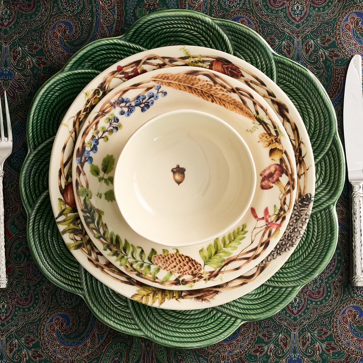 A stacked set of ceramic dinnerware including a small cream bowl, plates with a botanical and woodland print, and a large green rope-textured charger plate, arranged on a patterned tablecloth with cutlery partially visible at the sides.