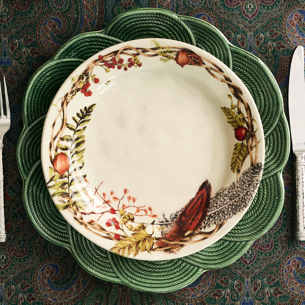 Moveable gif featuring a stacked set of ceramic dinnerware including a small cream bowl, plates with a botanical and woodland print, black plates, and a large green rope-textured charger plate, arranged on a patterned tablecloth with cutlery partially visible at the sides.