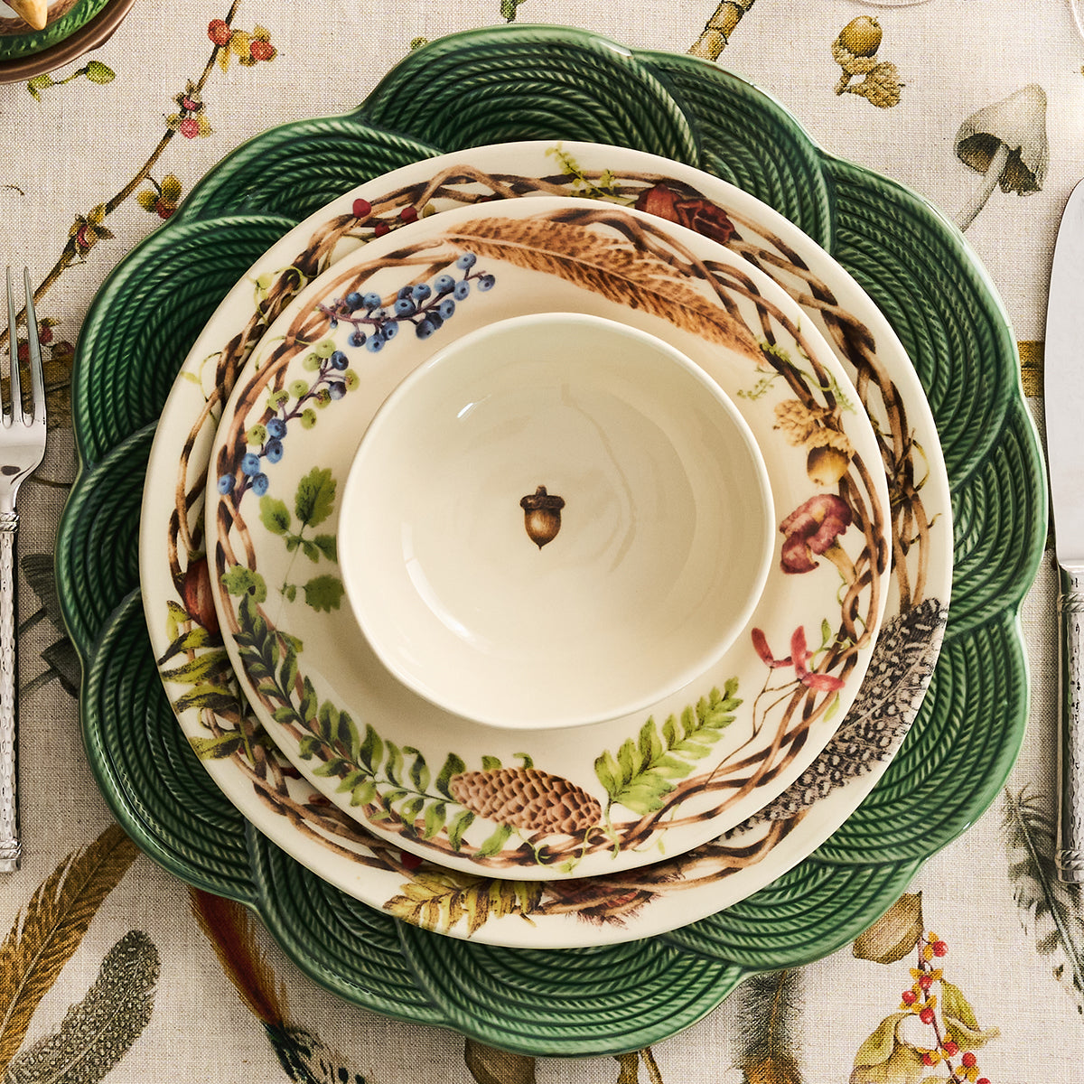 A stack of ceramic tableware plates and a small bowl with a woodland botanical print, placed on a green rope-textured placemat over a beige patterned tablecloth. Silver cutlery is partially visible at the left and right edges.