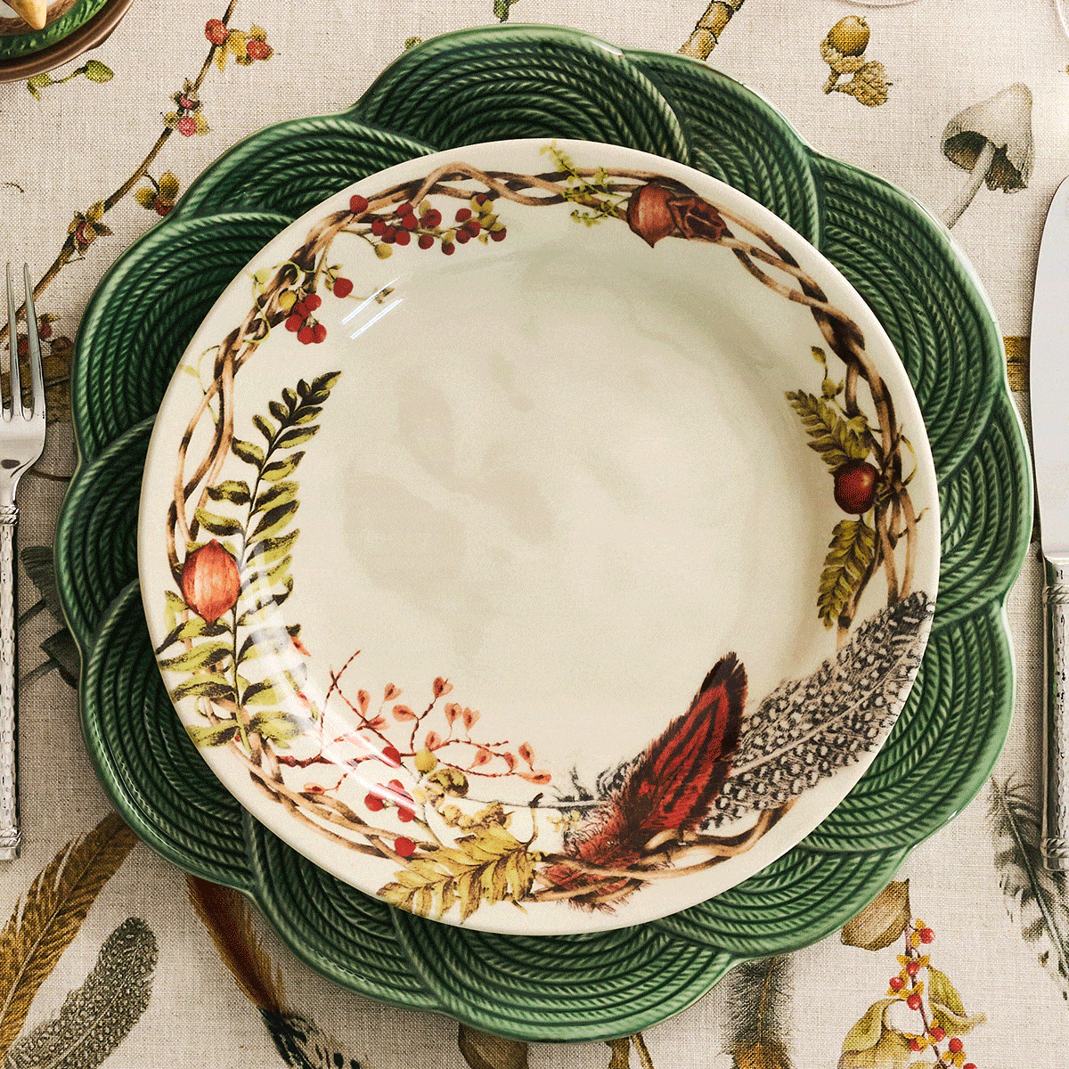 A festive table setting with a cream-colored napkin adorned with a leafy acorn napkin ring, silverware, and a decorative plate featuring a whimsical turkey motif and autumnal foliage on a nature-themed tablecloth.