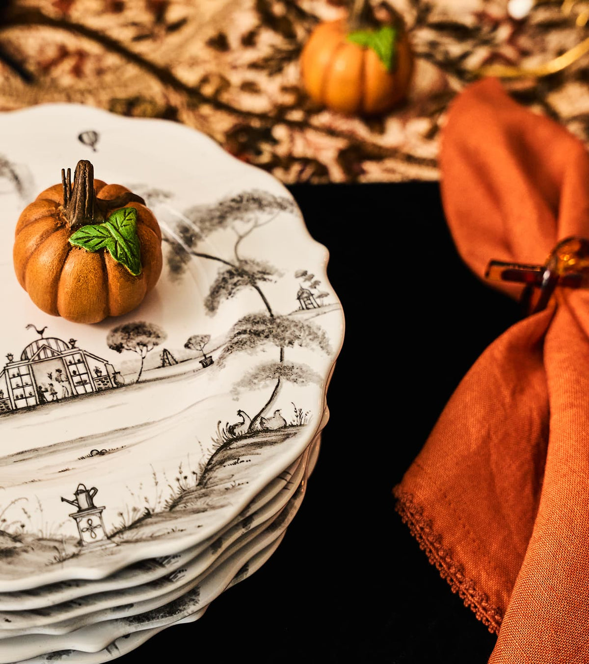 A stack of white ceramic plates with a black landscape toile de Jouy print, styled with small decorative pumpkins on a dark table next to an orange napkin.