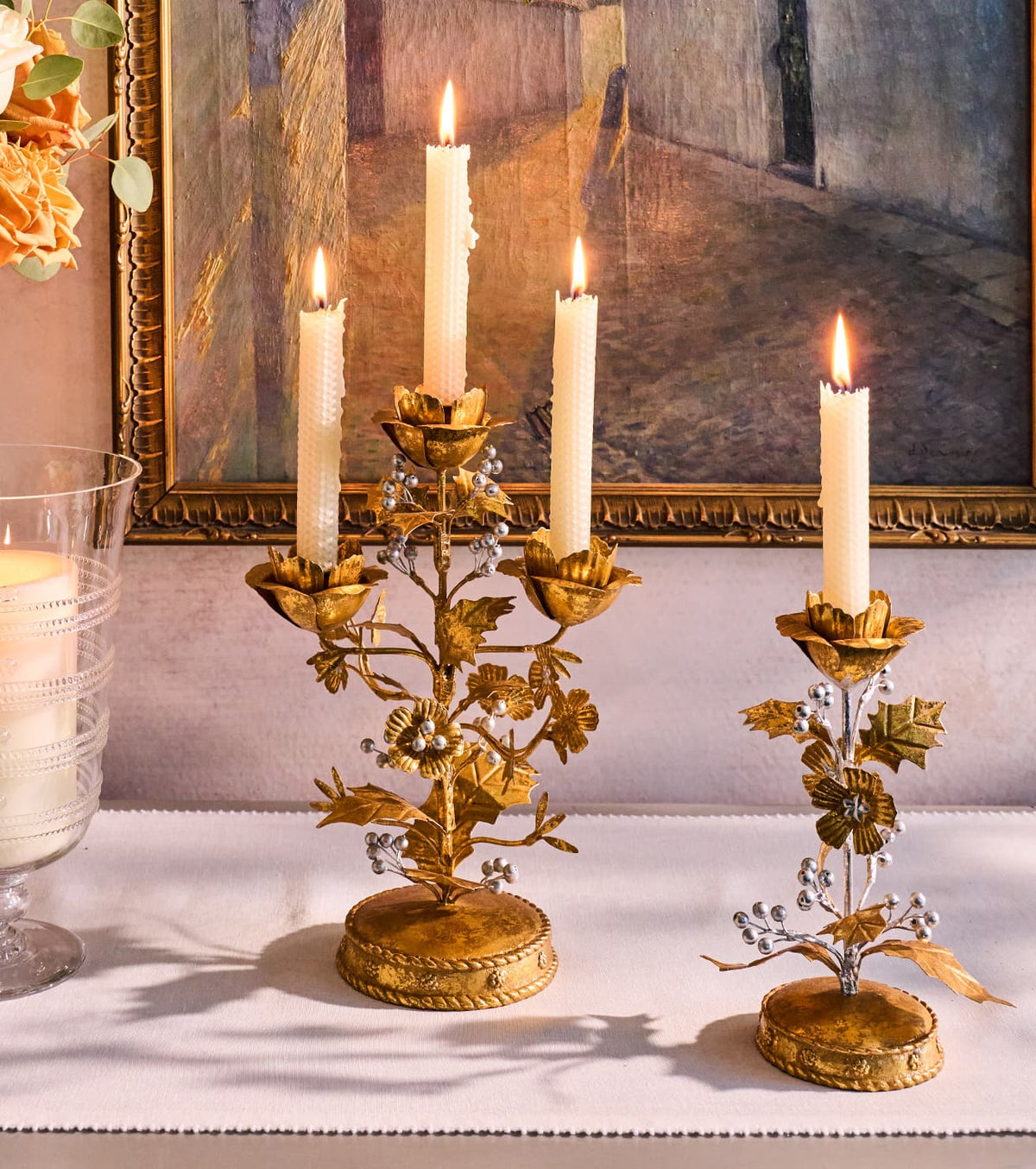 Two ornate gold metal candlestick holders with floral and leaf details, each holding a lit white candle, displayed on a white table runner in front of a framed painting.