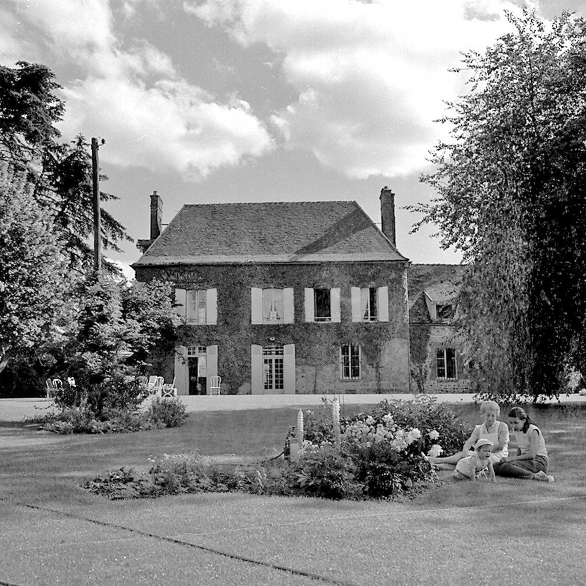 A black and white photograph shows a mother, child and baby sitting near the garden in front of a large country home in the farmlands of France.
